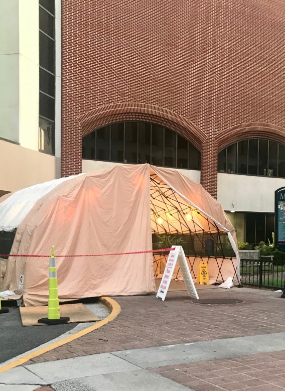 A large white tent with lights inside is shown outside a brick hospital building. A sandwich board sign advises that there is no visitation allowed due to COVID-19.
